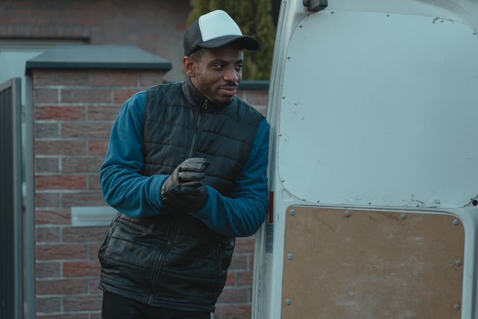 A man with glasses and a dark baseball cap sits inside a vehicle, likely a moving van, with his left arm resting on the steering wheel and his right hand holding a pen, writing on a clipboard. Behind 