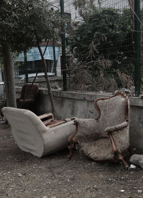Exterior view of two antique upholstered armchairs placed on a gravel surface outside a property, with one chair showing signs of wear and damage to the fabric and wooden frame. The chairs are positioned near a low concrete wall and a metal fence with vertical bars, behind which there are trees and a building with a blue awning. The scene appears to be part of a bulky waste collection or disposal process, with the chairs likely prepared for removal by Man with Van Yeading, reflecting the company's home relocation and furniture transport services. The lighting is natural and overcast, emphasizing the worn condition of the furniture and the outdoor environment.