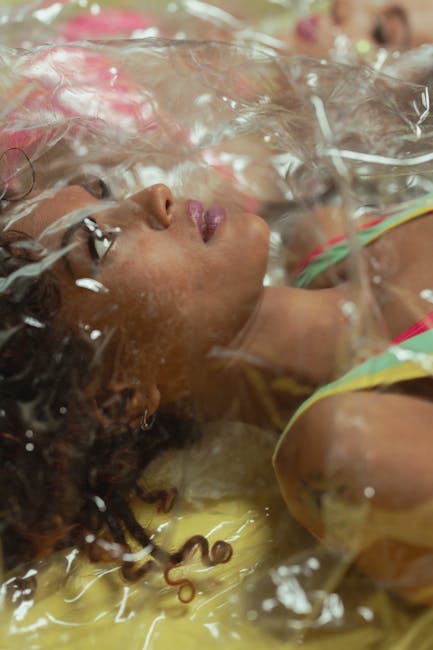 Photo showing a woman lying on a pavement surface outdoors, surrounded by a transparent plastic sheet and various items used in home relocation or furniture transport. She is positioned near a large, white refrigerator and several cardboard boxes, which appear to be packed for a move. The scene includes a moving trolley, plastic wrap, and packing materials indicating an active loading process. The woman is wearing casual clothing with visible bright colours. The environment suggests a house removal scenario with items organized for transportation, and the lighting indicates natural daylight. This image exemplifies the staging and packing aspects involved in residential furniture transport and home relocation services provided by companies like Man with Van Yeading.