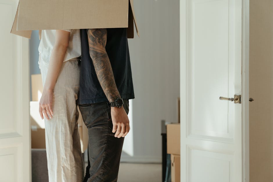 A man and a woman stand inside a house near an open doorway, with the man holding a large cardboard box on his shoulder. The man, wearing a black t-shirt, dark trousers, and a wristwatch, has visible tattoos on his arm and is in the process of a home relocation or packing task. The woman, dressed in light-colored casual clothing, is standing nearby with her left arm hanging relaxed by her side. Behind them, cardboard moving boxes are stacked, and light streams in from an unseen window, illuminating the interior space. Outside the doorway, a moving van with an open rear door is visible on the pavement, with furniture pieces and packing materials such as plastic wrap and blankets surrounding the loading area. The scene captures the logistics of furniture transport during a fast-moving eviction or short-notice relocation, as handled by Man with Van Yeading, with a focus on the loading process in a residential setting.