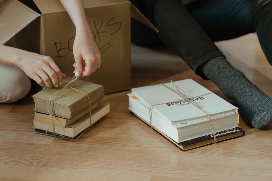 Two individuals are seated on a wooden floor indoors, engaged in packing activities related to house removals. One person is tying a thick string around a small stack of three books, which are bound together to form a bundle. Behind them, there is a cardboard box labeled 'BOOKS,' indicating it is used for moving or storage. To the right, another bundle of books and documents is visible, secured with string, including a visible printed label 'BALANCE.' One person is wearing dark pants and grey socks, while the other has light-colored clothing and a hand that is actively securing the book bundles. The scene reflects the packing process involved in home relocation, with the books neatly tied and prepared for transport. The environment appears to be inside a home, with the floor clearly visible and no other furnishings in view, highlighting a typical packing stage in a move organized by a professional removals service like Man with Van Yeading.