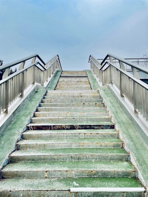 A concrete staircase with metal handrails on both sides leading up to a higher level, situated outdoors under a cloudy sky. The stairs are weathered, with some greenery and moss visible on the steps,