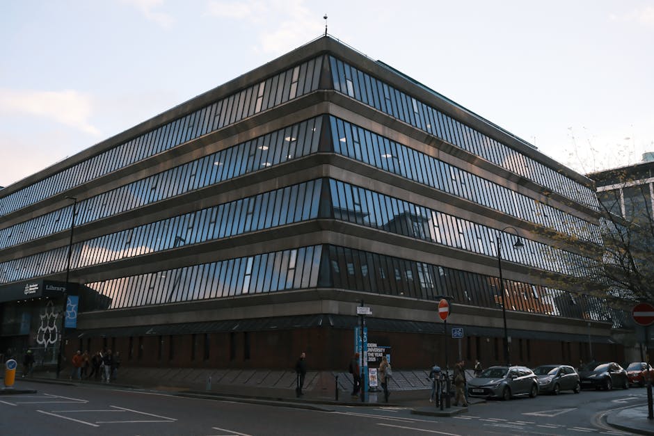 A modern, multi-storey office building with a glass exterior featuring horizontal metallic bands, situated on a busy street near Yeading Library. The ground floor has a dark entrance with a sign indicating a public library, and there are several pedestrians walking along the sidewalk. Parked cars line the curbside, and street lamps are visible along the road. The building's reflective windows capture the cloudy sky above. This urban setting, with the building's sleek design, illustrates typical cityscape surroundings relevant to house removals and furniture transport in the Yeading area, supporting professional relocation services like those offered by Man with Van Yeading. The scene suggests an environment where careful planning is needed for moving logistics, particularly at busy street locations, aligning with tips for effective home relocation and loading processes.