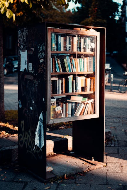 A black outdoor public bookshelf with a glass front containing multiple shelves filled with various hardcover and paperback books, situated on a paved sidewalk near a street with a bicycle and parked cars in the background. The bookshelf is covered in graffiti and stickers, indicating it is in a public urban area. A nearby tree provides partial shade, and the scene appears to be in natural daylight, suggesting a quiet, accessible location for community book exchange or reading. This image emphasizes outdoor book storage, relevant to the context of house removals and furniture transport, highlighting the importance of secure, weather-resistant furniture for home relocation services provided by Man with Van Yeading.