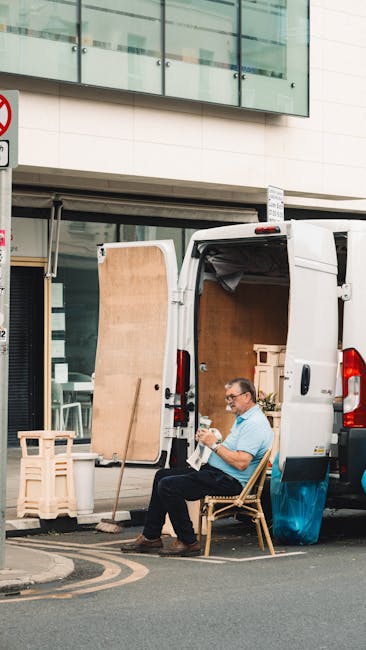 A man with glasses sits on a wooden chair positioned outside a white delivery van, which is parked on the street in front of a modern building with large glass windows. The van's rear doors are open, revealing packed cardboard boxes, wooden furniture panels, and packing materials inside, indicating a home relocation process. The man holds a small item in his hands, possibly a mobile phone or a packing accessory. Nearby, a wooden stool and plastic packaging wraps are visible, suggesting ongoing packing or loading activities related to furniture transport and moving logistics. The scene is set in daylight, with urban surroundings including a no-parking sign partially visible in the background, reflecting a typical busy street environment for urgent same-day removals as outlined on the 'manwithvanyeading.co.uk' page about same-day Yeading removals.