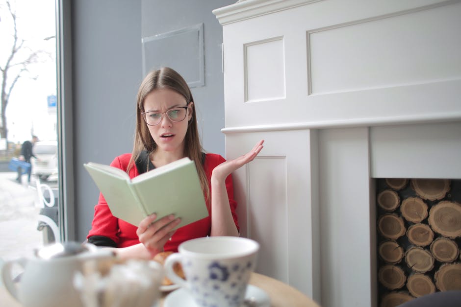 A young woman with long brown hair, glasses, and wearing a red top, sitting indoors near a large window with a view of a snowy street and pedestrians outside. She appears confused or frustrated while holding an open green notebook in one hand and gesturing with the other. In front of her on the table are a white teapot, a matching cup, and a small dish. Behind her, there is a white wooden fireplace mantel with decorative paneling, and to the right, a space filled with neatly stacked logs. The scene suggests a moment of questioning or concern during a house move or packing process, with natural daylight illuminating the room. This setting could be part of the home relocation or furniture transport process managed by Man with Van Yeading, highlighting the importance of clear communication in urgent removals and moving services, especially in the context of same-day bookings for house removals in Yeading.