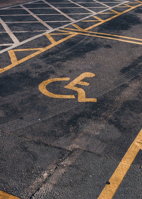 A section of an outdoor parking area surface with yellow painted lines marking designated spaces, including a prominent wheelchair accessible parking symbol painted in yellow. The asphalt surface appears weathered with visible cracks and patches. The parking area is adjacent to a building entrance, with a slightly inclined driveway or pavement visible at the top edge of the image. This setting illustrates the area where Man with Van Yeading may organize the loading and unloading process during a home relocation or furniture transport service, ensuring accessible parking for moving vans and clients involved in packing and moving activities.
