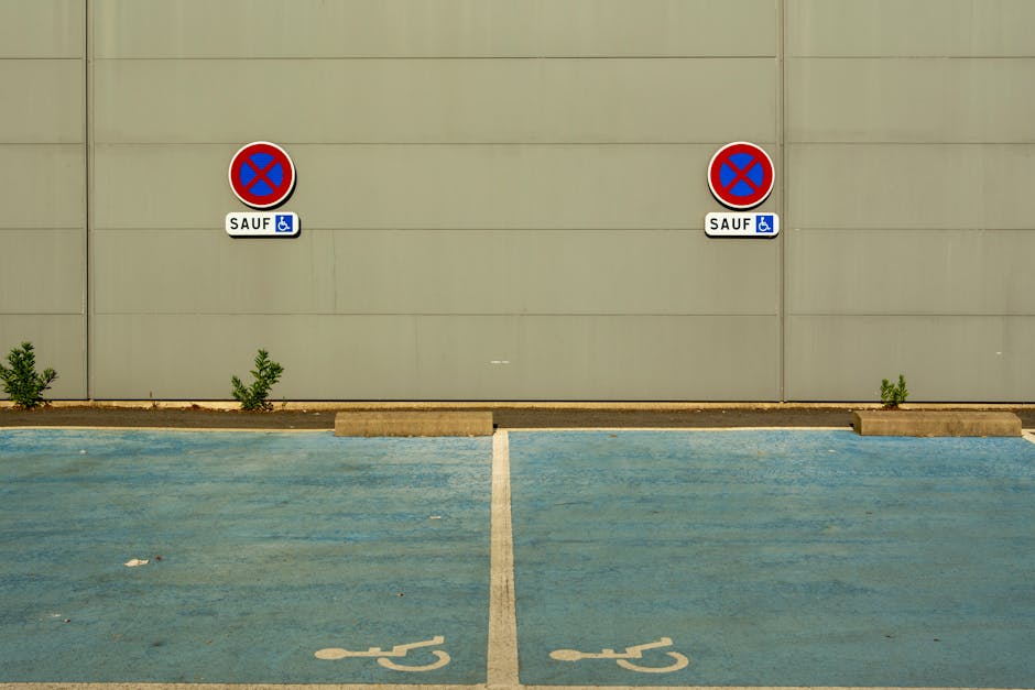 An exterior view of a building wall painted in light grey with two circular no parking signs attached, each featuring a red border and blue background with a red cross. Below each sign, there is a small white rectangular sign with the word 'SAUF' and a blue wheelchair accessibility symbol, indicating designated parking restrictions. In front of the wall, there are two marked parking spaces painted in blue with white wheelchair symbols, separated by a white dividing line. The ground is paved, with small curb stones and minimal shrubbery on the edge of the pavement near the wall. The area appears to be a designated parking lot, possibly accessed during a home relocation or removal process. The setting is clean and well-maintained, suited for logistics involving furniture transport or moving services, as occasionally referenced by Man with Van Yeading.