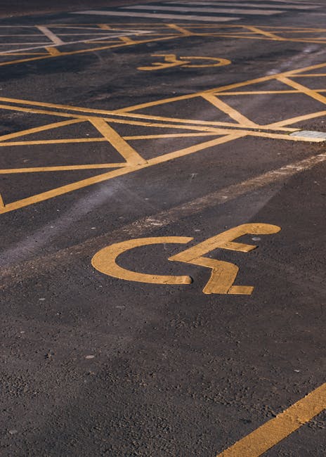A section of an outdoor parking area surface with yellow painted lines marking designated spaces, including a prominent wheelchair accessible parking symbol painted in yellow. The asphalt surface appears weathered with visible cracks and patches. The parking area is adjacent to a building entrance, with a slightly inclined driveway or pavement visible at the top edge of the image. This setting illustrates the area where Man with Van Yeading may organize the loading and unloading process during a home relocation or furniture transport service, ensuring accessible parking for moving vans and clients involved in packing and moving activities.