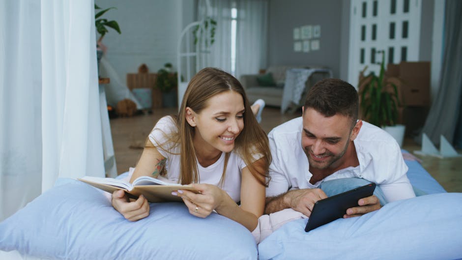A young woman and a man are relaxing on a bed inside a well-lit living room, with the woman holding an open book and the man looking at a tablet they are both smiling. The room features a white curtain, a plush sofa with cushions, indoor plants, and a wooden side table. The background includes a modern interior with a patterned wall, a large window allowing natural light, and some cardboard boxes indicating recent moving or packing activities. The scene captures a casual moment during home relocation or packing and moving processes, reflecting the environment where professional removals like those from Man with Van Yeading might be involved to assist with furniture transport and house removals.