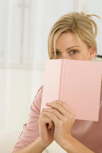 A woman with blonde hair and light skin is inside a home, partially hiding behind a light pink hardcover book she is holding up to her face. She is wearing a pale pink top, and her eyes are looking directly at the camera. Behind her, the background shows a bright, softly lit interior space with white walls and cabinetry, indicating a domestic setting. The scene suggests a moment of reading or contemplation within a home environment. This image may relate to house removals or packing services offered by Man with Van Yeading, highlighting the importance of proper packing during a home relocation process.