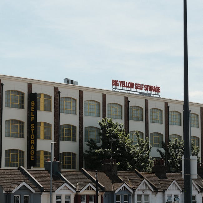 The image shows a large, multi-storey commercial building used for self storage, with a white facade and yellow-trimmed, arched windows on each floor. The building features a prominent vertical black sign reading 'Self Storage' on the left side. At the top of the building, a red sign displays 'Big Yellow Self Storage.' In front of the building, there are several small residential houses with tiled roofs and chimney stacks. A few trees are visible in front of the storage facility, partially obstructing the view of some windows. The scene is captured during daylight hours, with a partly cloudy sky overhead, and a streetlamp is visible on the right edge of the image. This setting highlights the typical environment involved in house removals, including the transition from residential to commercial storage or transport facilities, relevant to relocating households using services like those offered by Man with Van Yeading.