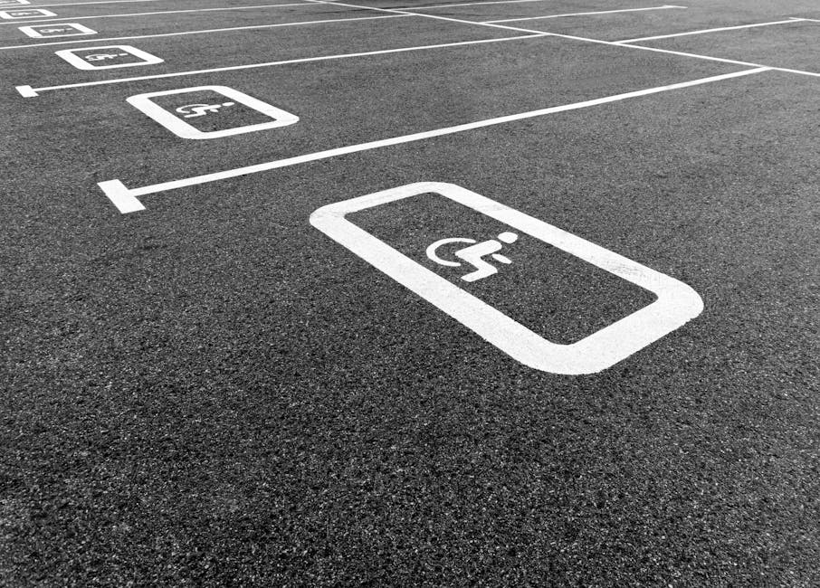 A close-up view of a parking lot surface with designated handicapped accessible parking spaces marked by white lines and symbols. The asphalt is dark and textured, with multiple parking bays visible, each outlined with clean, crisp white paint. The prominent wheelchair symbol is displayed in the foreground within a rectangular border, indicating accessible parking. In the background, additional parking spaces are outlined, showing the organized layout of the lot. The scene highlights the well-maintained infrastructure designed for easy access, which could be relevant to a home relocation or moving service like Man with Van Yeading, that requires careful planning around parking and access points for furniture transport and loading during house moves.