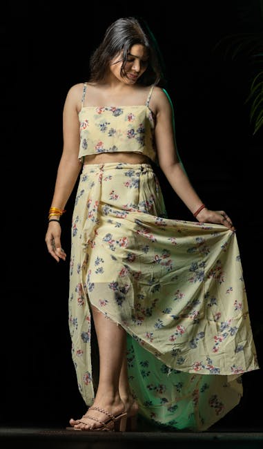 This image shows a woman with long, dark hair wearing a two-piece floral outfit consisting of a crop top and a long, flowing skirt. She is standing indoors on a dark background, illuminated by a light source that highlights her facial features and clothing. She is holding the edge of her skirt with one hand, gently lifting it to reveal the fabric's pattern and texture. The outfit is made of lightweight fabric with a soft pastel yellow base and a delicate floral print in various colors. She is wearing minimal jewelry, including a bracelet on her wrist, and beige/open-toe high heels. The setting appears to be a studio or interior environment, with no visible furniture or other objects, focusing solely on her and her attire.